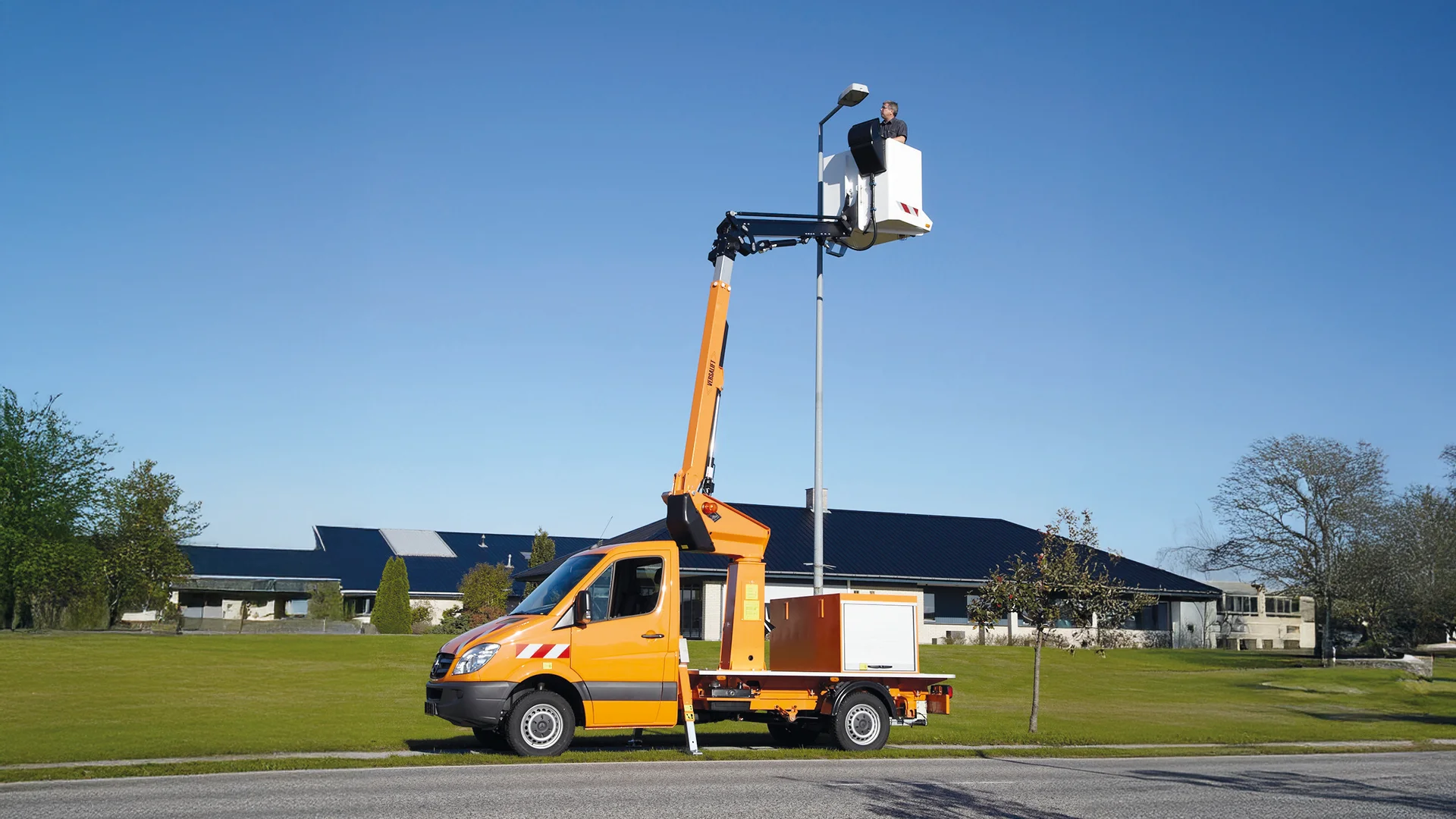 Seitliche Ansicht einer ausgefahrenen gelben Arbeitsbühne mit einer Person darauf, blauer Himmel im Hintergrund.