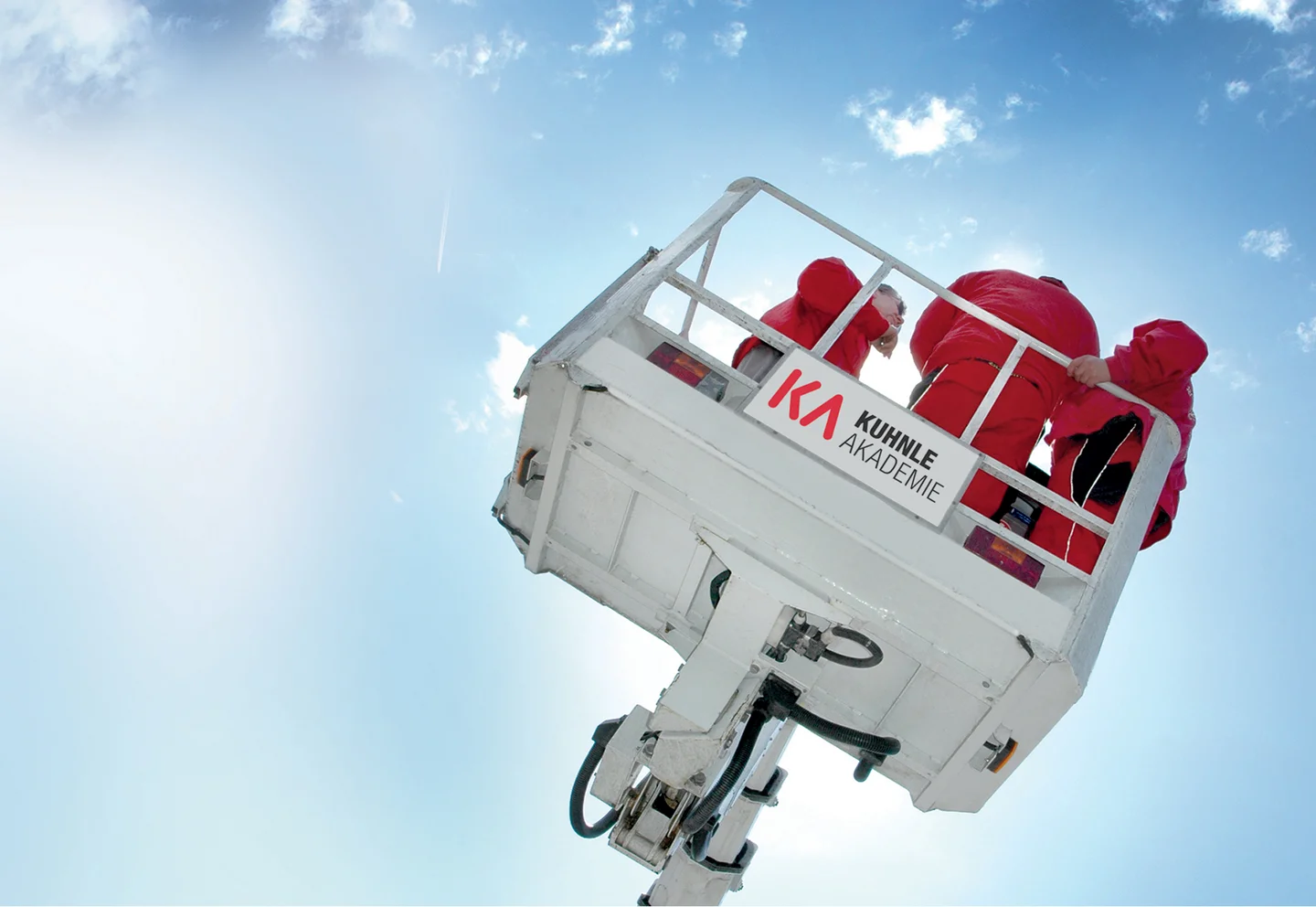 Arbeitsbühne in ausgefahrenem Zustand, darauf drei Personen in Rot, vor leicht wolkigem blauen Himmel.