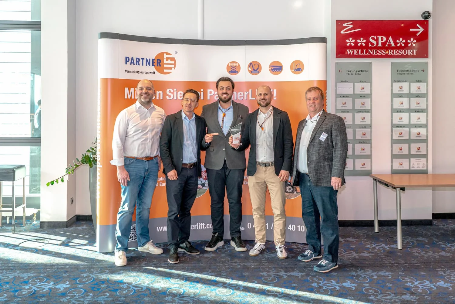 Fünf Männer vor Press Wall in einem Raum, links Fenster, rechts Tisch.