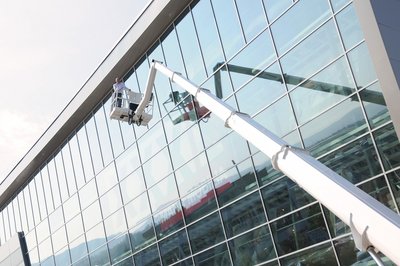Ausgefahrene Arbeitsbühne vor hohem gläsernem Gebäude, darauf eine Person, links oben Himmel und Wolken.