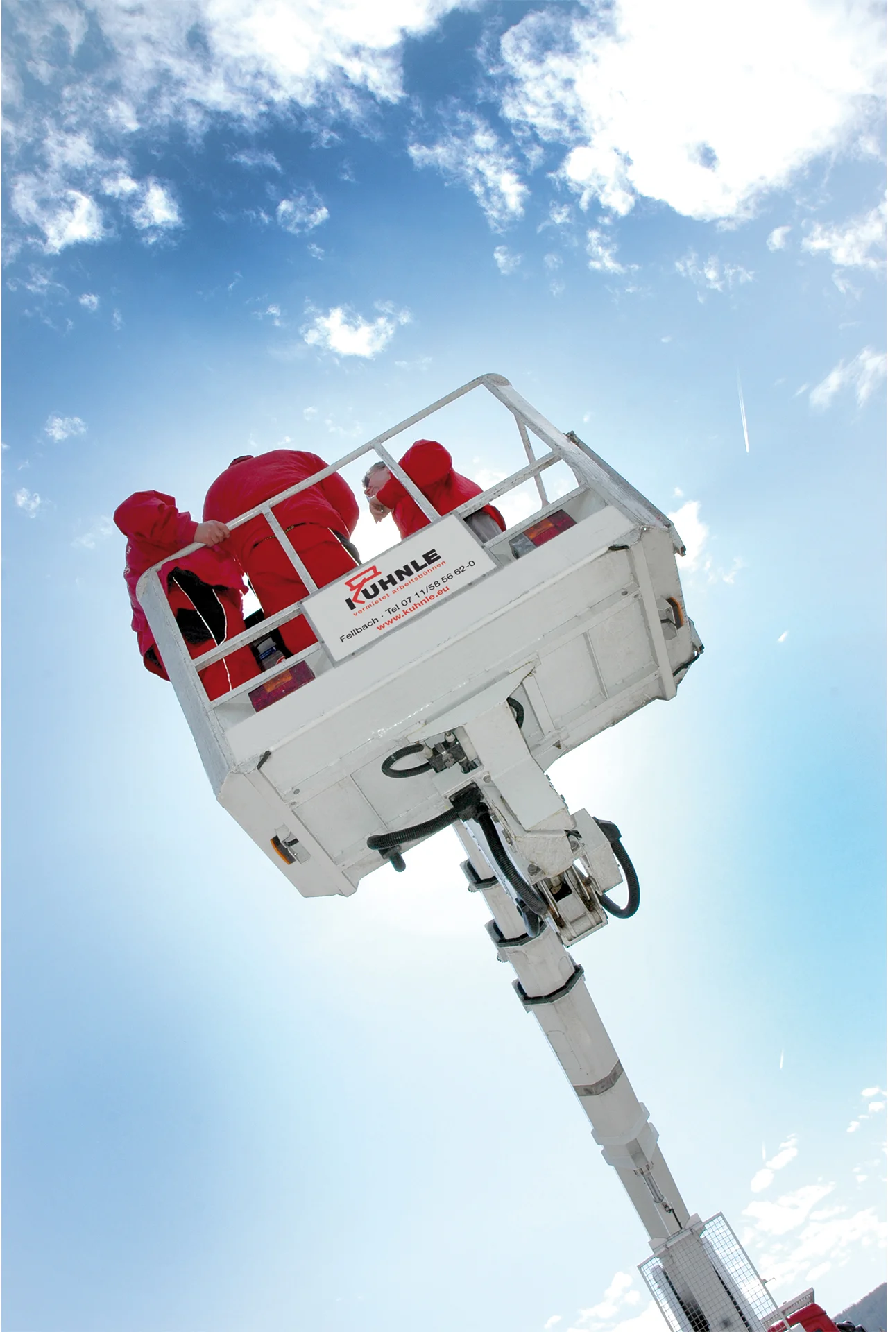 Arbeitsbühne in ausgefahrenem Zustand, darauf drei Personen in Rot, vor leicht wolkigem blauen Himmel.