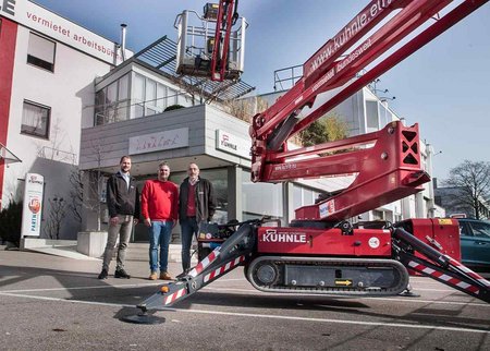 Gebäude, davor drei Personen, rechts rote Arbeitsbühne.