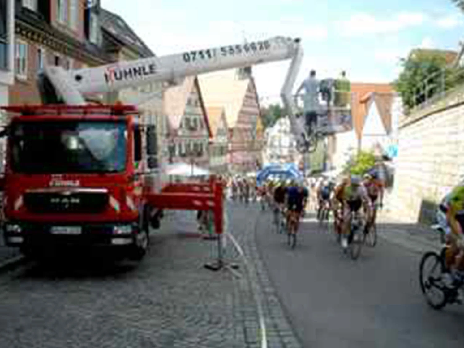 Gasse mit Häusern, links eine ausgefahrene Arbeitsbühne, rechts fahrradfahrende Personen.